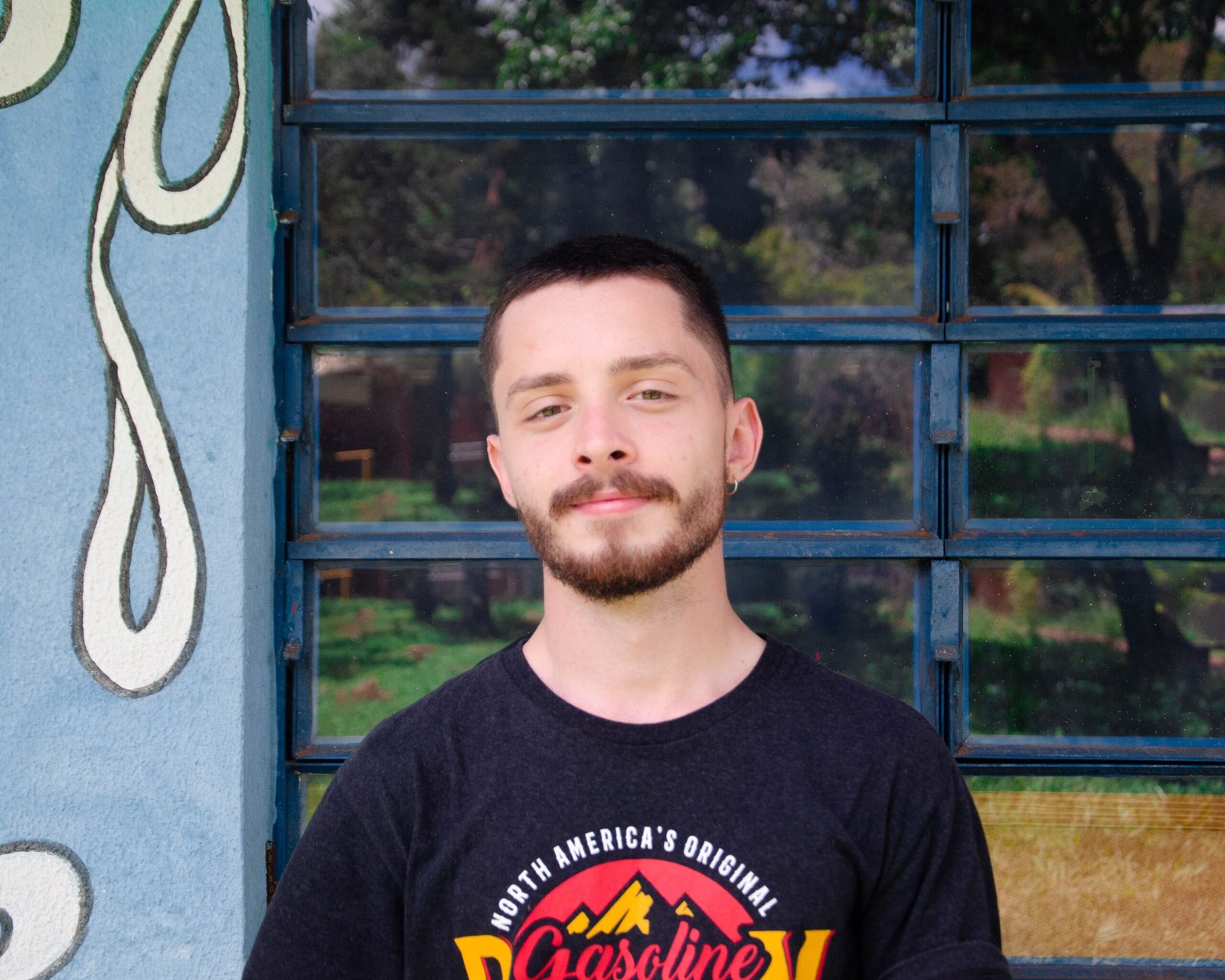 A man poses casually in front of a window, wearing a graphic tee.